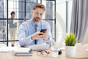 Handsome businessman in office using smartphone with collegues on the background.