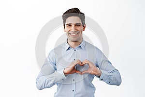 Handsome businessman showing heart sign and smiling, I love you like gesture, standing over white background in office