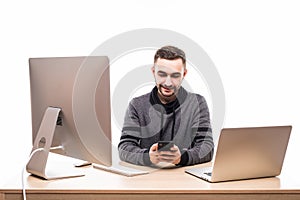 Handsome businessman sitting behind his desk with laptop and pc and typing on mobile phone isolated on white background
