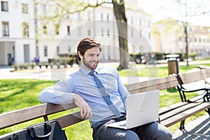 Handsome businessman sitting on city bench, typing on laptop. Working remotely from city park, waiting for business