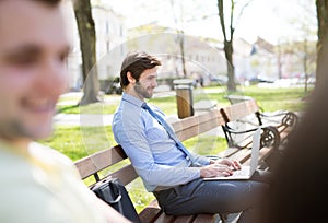 Handsome businessman sitting on city bench, typing on laptop. Working remotely from city park, waiting for business