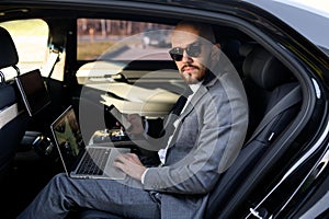 Handsome businessman sitting with laptop on the backseat of the car. travelling by a car and working on laptop computer
