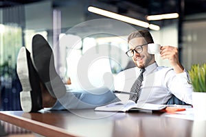 Handsome businessman sitting with legs on table and drinking coffee in office