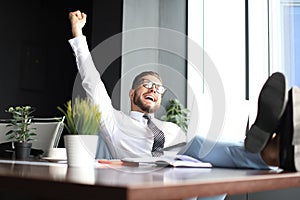 Handsome businessman sitting with legs on table and keeping arm raised and expressing joyful in office