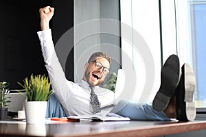 Handsome businessman sitting with legs on table and keeping arm raised and expressing joyful in office