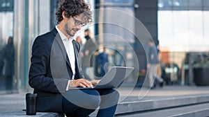 Handsome Businessman in a Suit is Sitting on Steps next to Business Center and Working on a Laptop