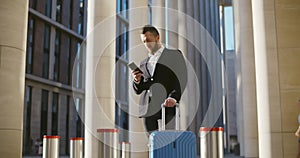 Handsome businessman in suit standing with suitcase outside airport terminal and using mobile phone