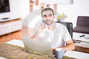 Handsome businessman is talking on the mobile phone and smiling while using a laptop in kitchen