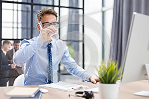 Handsome businessman using PC and drinking water in office area with collegues on the background