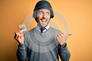 Handsome businessman wearing military helmet holding reminder paper with war message pointing and showing with thumb up to the