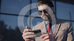 Handsome Bussinessman Wearing Sunglasses and Stylish Suit with Tie Holding his Smartphone Looking into it front of