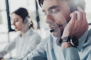 handsome call center manager in headphones with mike sitting at workplace while his colleague sitting