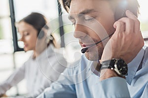 handsome call center manager in headphones with mike sitting at workplace while his colleague sitting