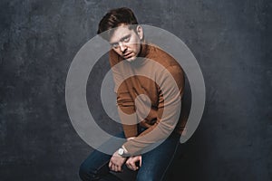 Handsome casual man sits on chair near a dark gray wall and looks to side, portrait picture.