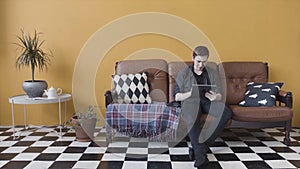 Handsome casual young man using a tablet at home sitting on a sofa in his living room, browsing the internet. Stock