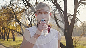 Handsome Caucasian athletic man in white singlet standing in sunlight and drinking water. Young sportive boy training