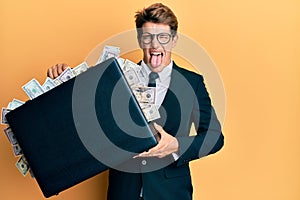 Handsome caucasian business man holding briefcase full of dollars sticking tongue out happy with funny expression