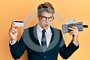 Handsome caucasian business man holding credit card and wallet with dollars in shock face, looking skeptical and sarcastic,