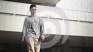 Handsome Caucasian cool man in sunglasses is walking along city street. Young stylish guy on blurred background urban architecture