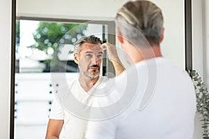 Handsome Caucasian grey-haired man with beard looks in the mirror healthy skin at home. Men's personal care. Middle aged man