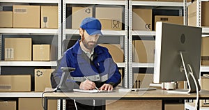 Handsome Caucasian mailman in blue iniform and cap sitting at table in postal office store and working at computer