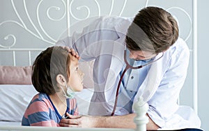 Handsome caucasian male doctor wearing white gown touching little boy`s head getting sick, measure fever and using stethoscope to