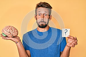 Handsome caucasian man with beard holding brain and question mark puffing cheeks with funny face