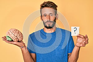 Handsome caucasian man with beard holding brain and question mark relaxed with serious expression on face