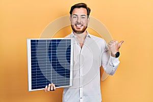 Handsome caucasian man with beard holding photovoltaic solar panel pointing thumb up to the side smiling happy with open mouth
