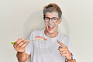 Handsome caucasian man eating prawn sushi using chopsticks smiling happy pointing with hand and finger