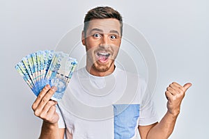Handsome caucasian man holding south african rand banknotes pointing thumb up to the side smiling happy with open mouth