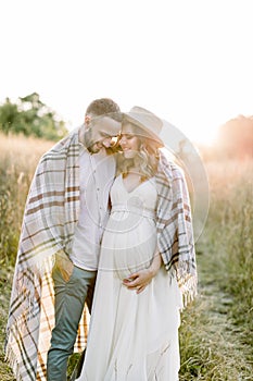 Handsome Caucasian man hugging his attractive pregnant woman in straw hat and white dress in sunny day in field. Happy