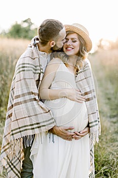 Handsome Caucasian man hugging his attractive pregnant woman in straw hat and white dress in sunny day in field. Happy