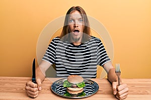 Handsome caucasian man with long hair eating a tasty classic burger afraid and shocked with surprise and amazed expression, fear