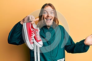 Handsome caucasian man with long hair holding red casual shoes celebrating achievement with happy smile and winner expression with