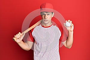 Handsome caucasian man playing baseball holding bat and ball thinking attitude and sober expression looking self confident