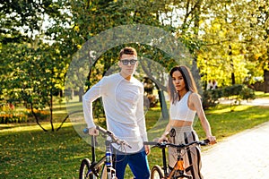 Handsome caucasian man and pretty hispanic woman are standing in the park, holding their bikes, posing for the photo and looking