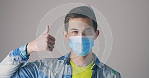 Handsome caucasian man in protective medical mask shows thumbs up, like gesture. Happy guy on gray background. Winner