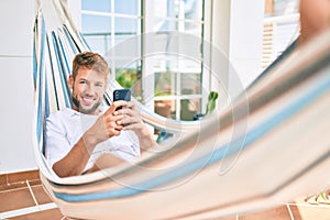 Handsome caucasian man smiling happy resting on a hammock at the terrace using smartphone