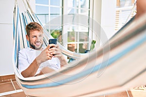 Handsome caucasian man smiling happy resting on a hammock at the terrace using smartphone