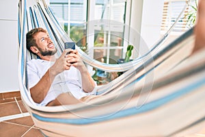 Handsome caucasian man smiling happy resting on a hammock at the terrace using smartphone