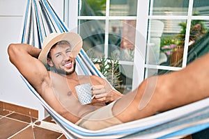 Handsome caucasian man smiling happy resting on a hammock at the terrace wearing summer hat drinking a cup of coffee