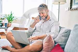 Handsome caucasian man smiling happy sitting on the sofa using computer laptop speaking on the phone