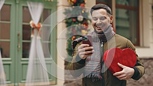 Handsome caucasian man standing in the winter decorating street holding heart shape gift and using smartphone. Handsome