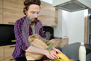 Handsome Caucasian man unpacking eco cardboard bag with healthy raw vegan food and a whole grain baguette, bananas and putting