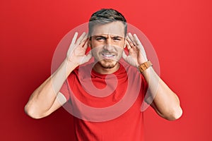 Handsome caucasian man wearing casual red tshirt trying to hear both hands on ear gesture, curious for gossip