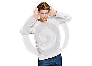 Handsome caucasian man wearing casual white sweater trying to hear both hands on ear gesture, curious for gossip