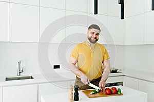 Handsome caucasian man in yellow cutting vegetables for meal or salad in modern white kitchen smiling