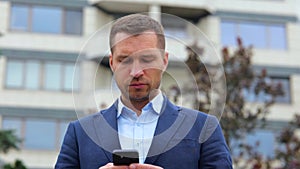 Handsome Caucasian stylish man with serious face standing in street in town texting on smartphone. Young successful