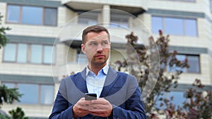 Handsome Caucasian stylish man with serious face standing in street in town texting on smartphone. Young successful